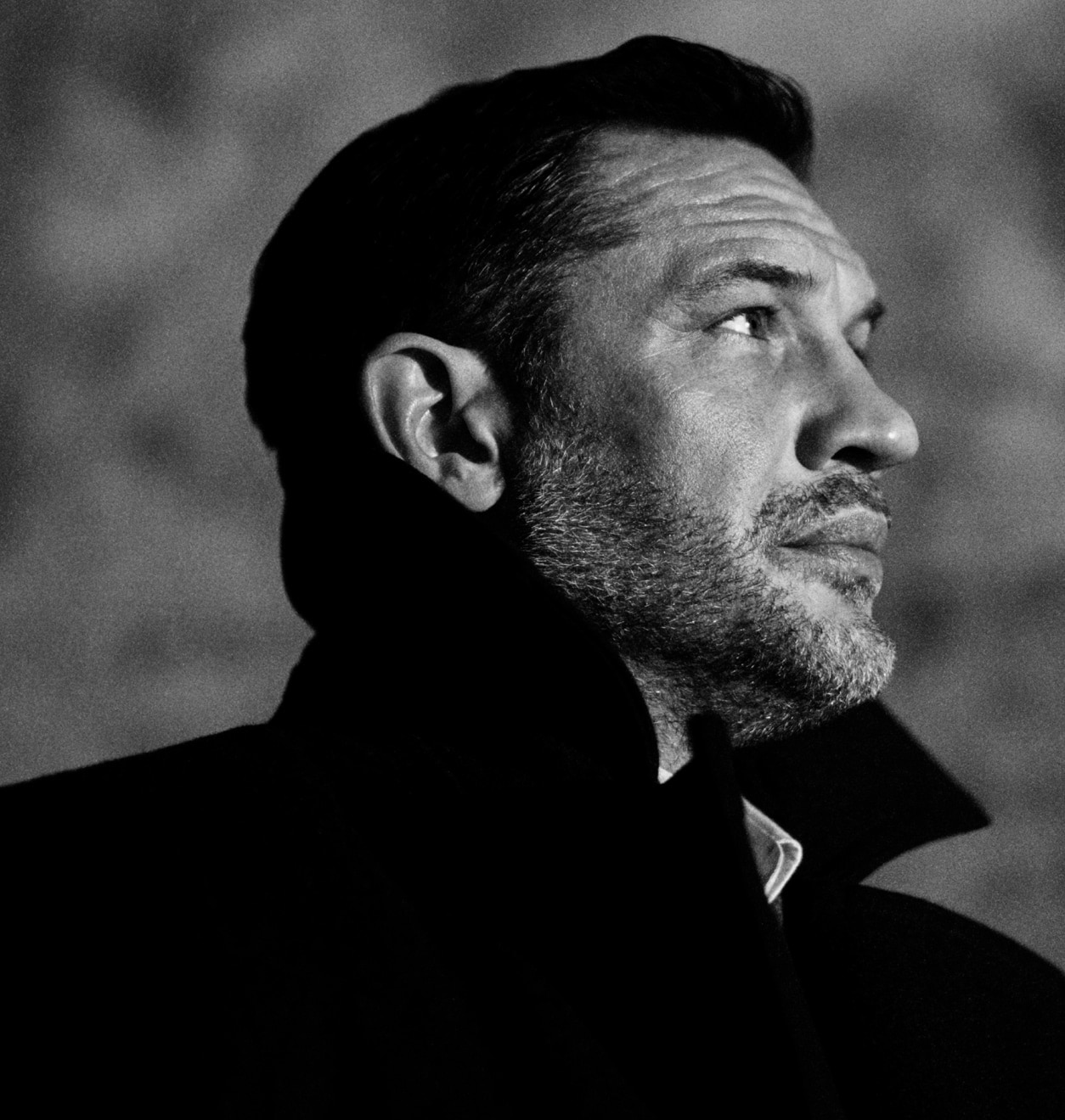 A side portrait of Tom Hardy in black and white, gazing into the distance. He wears a formal coat, with an upturned collar