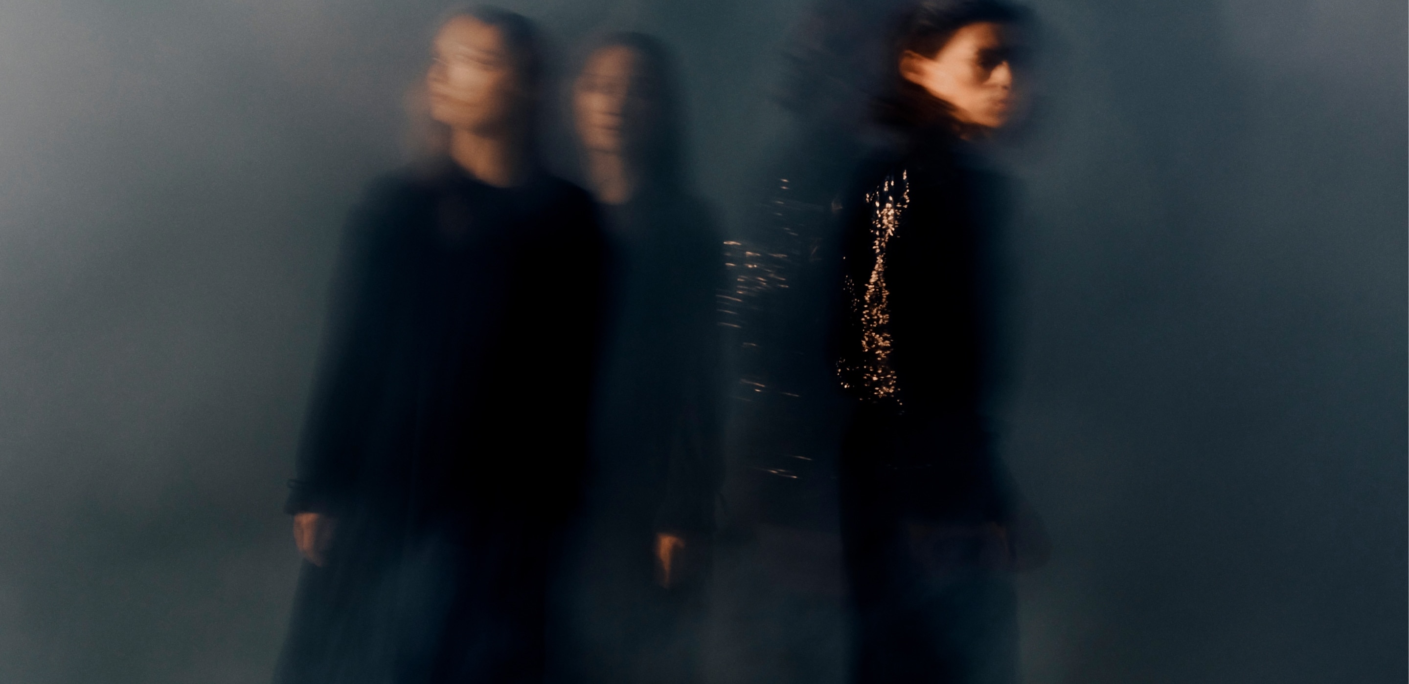 A few expressionless, blurry figures stand amongst each other against a dark backdrop. 