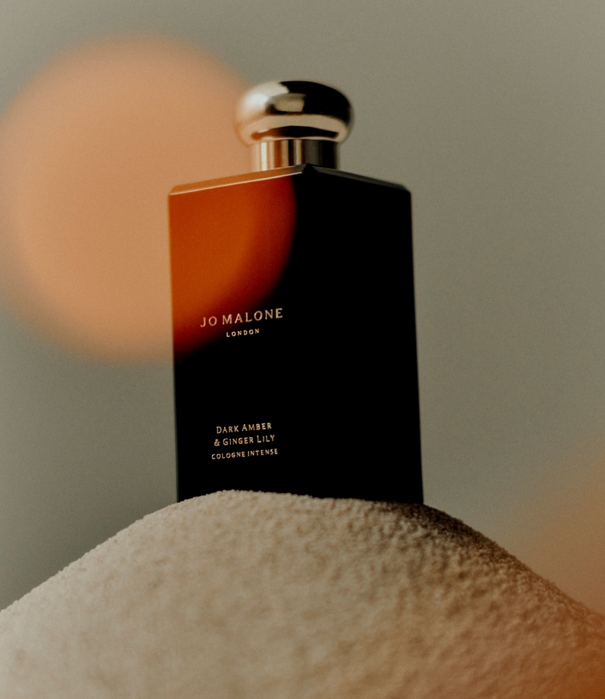 A Jo Malone London cologne, in a dark bottle, rests on a small mound of sand against a dark but neutral backdrop. 