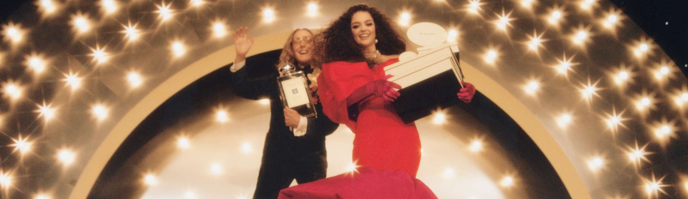 Woman in red dress holding jo malone gift boxes on a gold glittering stage