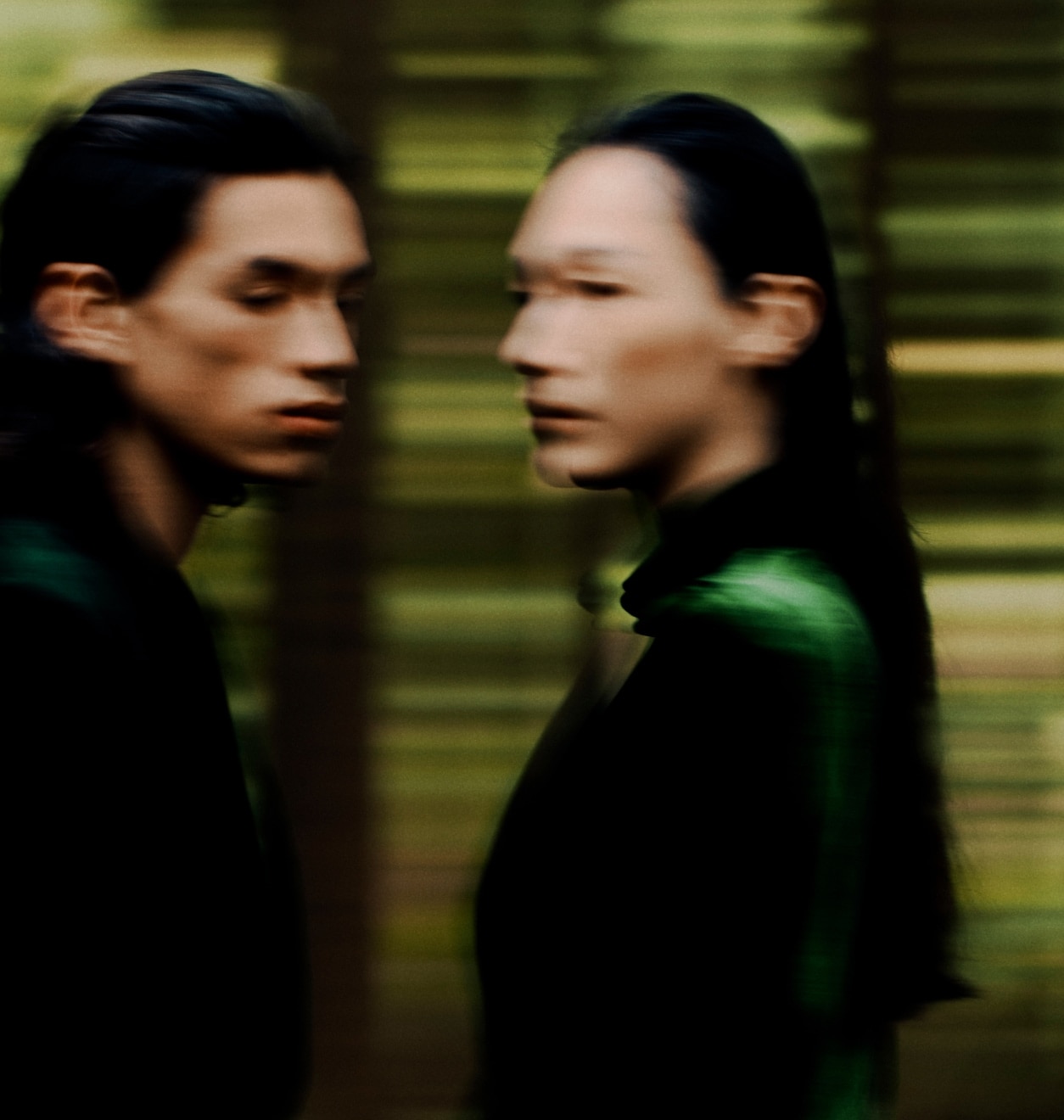 Two figures in green stand expressionless, facing each other against a dark, blurred backdrop of trees. 