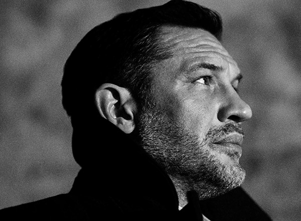 A side portrait of Tom Hardy in black and white, gazing into the distance. He wears a formal coat, with an upturned collar.