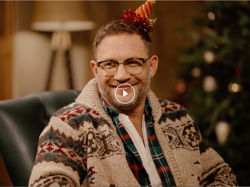 A clapperboard reveals Tom Hardy sat next to his father, against a light backdrop. He is wearing a grey jumper and glasses.