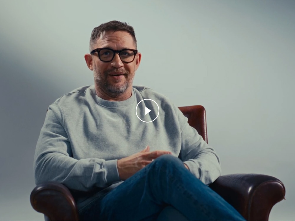 A clapperboard reveals Tom Hardy sat on a dark chair, against a light backdrop. He is wearing a grey jumper and glasses.