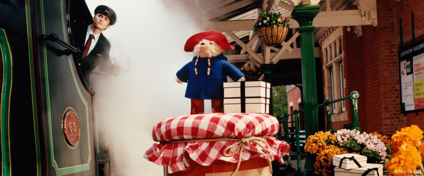 Paddington bear beside cream boxes on a giant Orange Marmalade jar surrounded by a train conductor extending from a steamy train